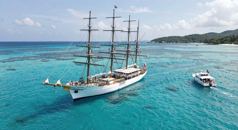 A four masted sailing cruise ship at anchor in a turquoise bay with a small tender approaching.