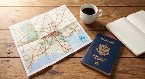 A folded paper map and a passport lying on a wooden table beside a small cup of coffee and an open notebook.