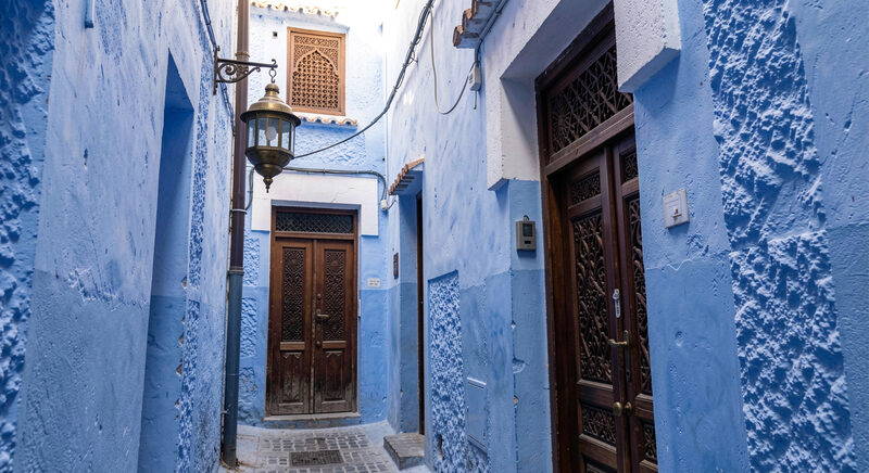 A narrow blue painted alley in a Moroccan medina with a hanging brass lantern and a carved wooden door.